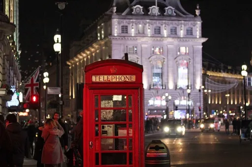A red telephone box stands in the foreground, with a busy street and illuminated buildings in the background at night.
