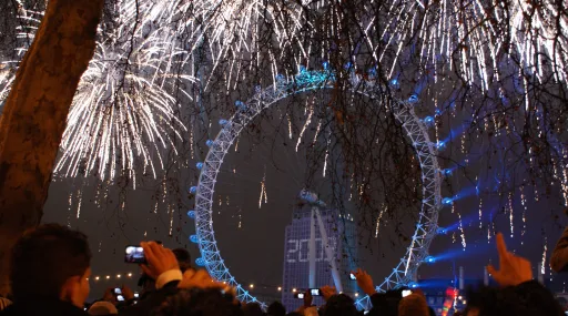 Fireworks illuminate the night sky above the London Eye, with people holding cameras in the foreground.
