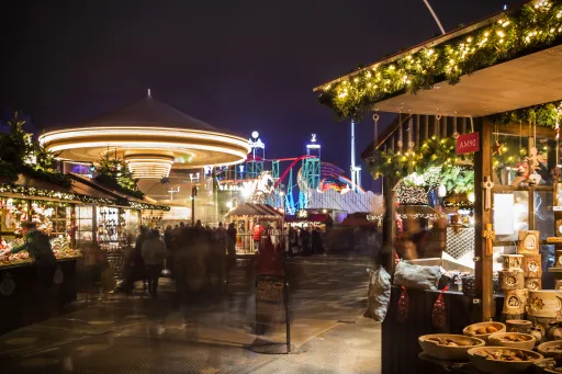 A festive market scene at night with illuminated stalls, a carousel, and a roller coaster in the background.