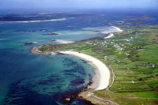 Aerial view of a coastal area featuring a sandy beach, green fields, and blue waters with rocky outcrops.