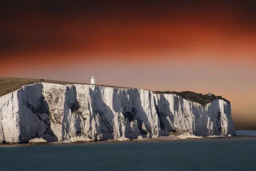 White lighthouse stands on top of a steep white cliff against a colourful sky at sunset.
