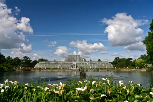 A large glass conservatory beside a lake, surrounded by greenery and flowers, under a blue sky with clouds.