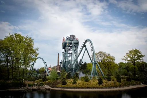 Roller coaster with riders is seen against a backdrop of trees and a cloudy sky.