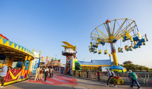 Vibrant amusement park scene with rides, colourful structures, and people walking along a path.