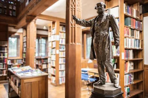 Bronze statue stands in a library surrounded by bookshelves and wooden furnishings.