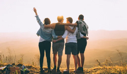 Four people stand together on a hilltop, facing a sunset with arms around each other.