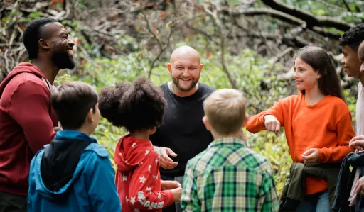 Group of people are gathered in a forest, smiling and engaging in conversation.