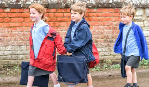 Three children walk along a path, wearing jackets and carrying bags, with a brick wall in the background.