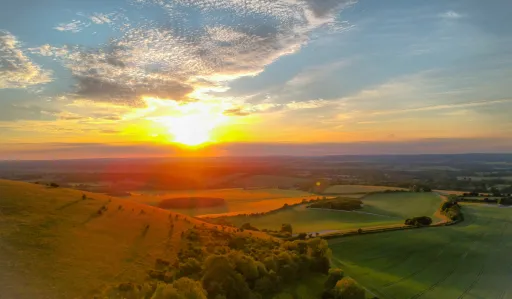 Sunset over rolling hills and fields, with a vibrant sky and scattered clouds.