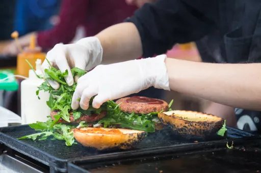 A person wearing gloves adds fresh greens to a burger on a grill, with toasted bun halves nearby.