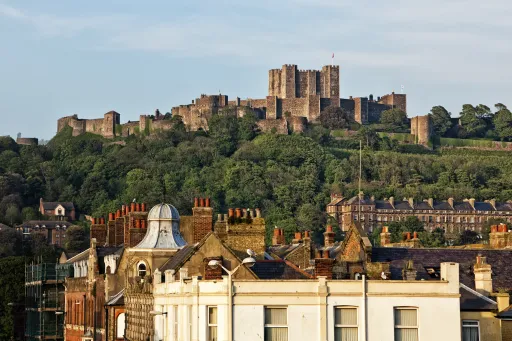 A historic castle sits atop a green hill, overlooking a row of buildings with various roof styles in the foreground.