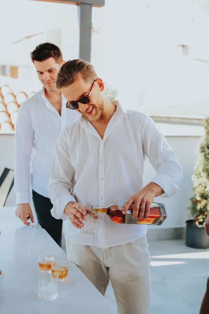 Friends enjoying drinks on a sunny outdoor terrace, with a man in a white shirt pouring whisky into a glass during a social gathering.