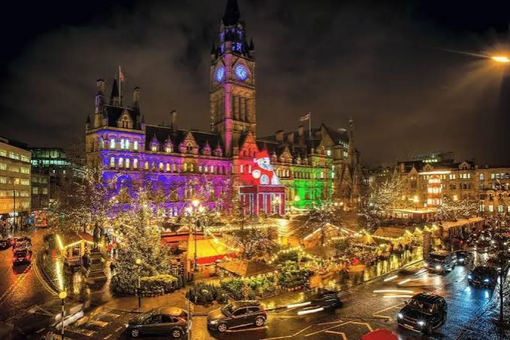 Manchester Christmas market in Albert Square at night, with illuminated stalls and festive lights outside Manchester Town Hall.
