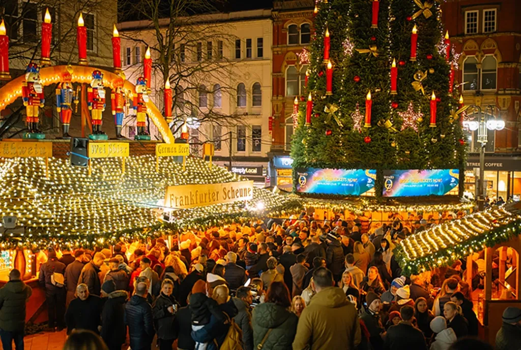Crowds at Birmingham Frankfurt Christmas Market, with illuminated wooden stalls and a large decorated Christmas tree at night.