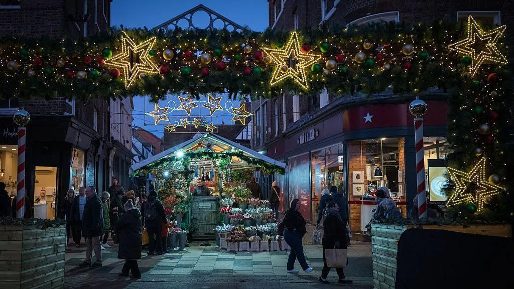 Visitors browsing a Christmas market stall beneath festive garlands, colourful baubles and illuminated stars at dusk.
