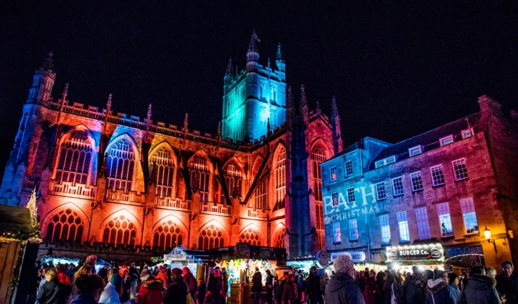 Bath Christmas Market at night, with visitors browsing festive stalls beneath Bath Abbey illuminated in colourful lights.