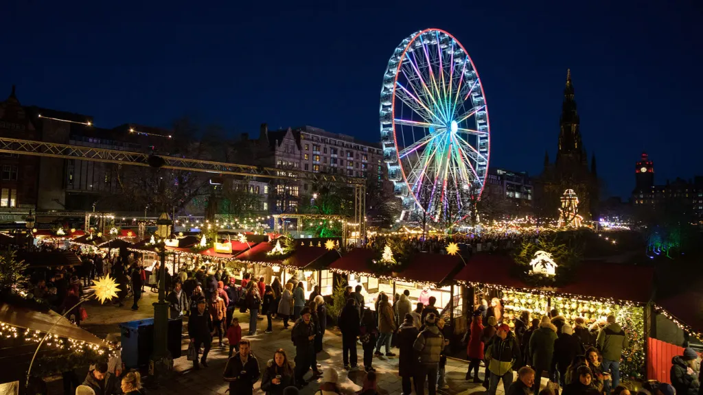 Edinburgh Christmas Market at night, with illuminated wooden stalls, crowds and a colourful Ferris wheel beside the Scott Monument.