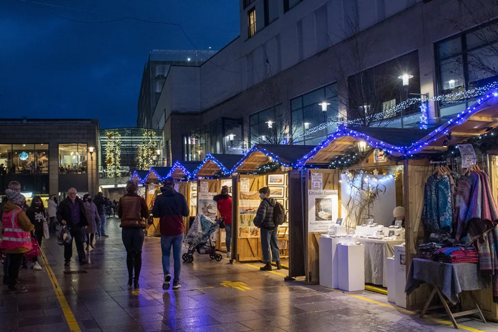  Visitors browsing wooden Christmas market stalls decorated with blue fairy lights, selling jewellery, clothing and crafts at dusk.