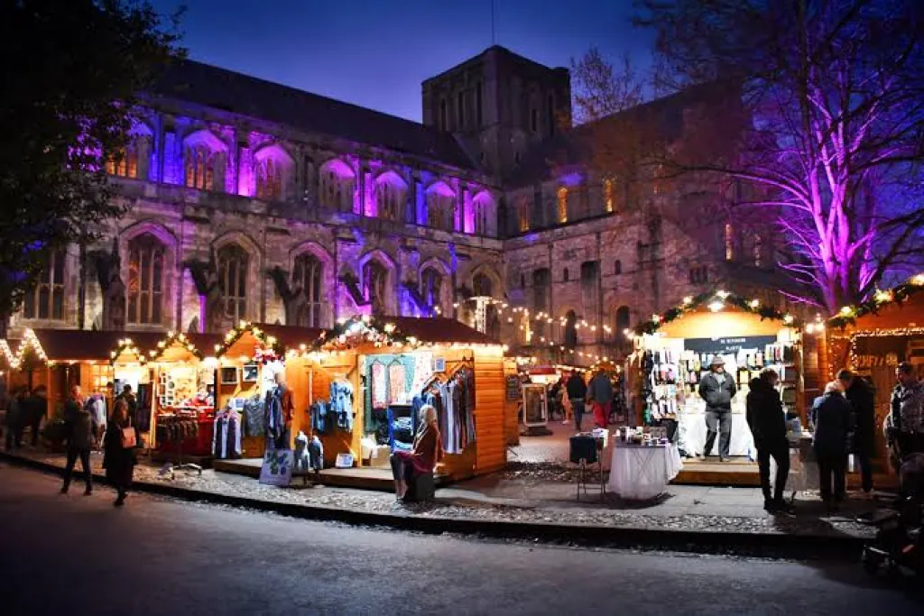 Winchester Cathedral Christmas Market at dusk, with wooden stalls and visitors beneath cathedral buildings illuminated in purple.
