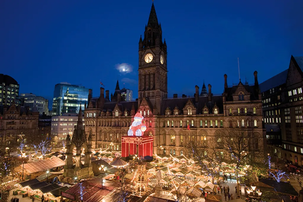 Manchester Christmas Market in Albert Square at dusk, with illuminated stalls and a large Santa decoration outside Manchester Town Hall.