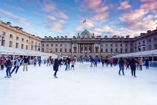 People ice skating on a rink in front of a large historic building under a blue sky with clouds.