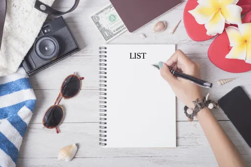 Hand writes LIST on a blank notebook surrounded by a camera, sunglasses, and beach items.