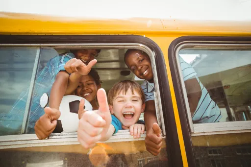 Four children are smiling and giving thumbs up from a school bus window, with a soccer ball visible inside.