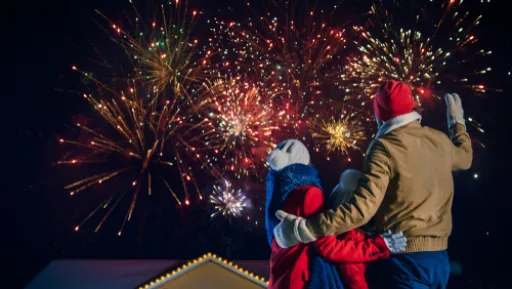 Three people watch fireworks in the night sky, standing together in front of a house with lights.
