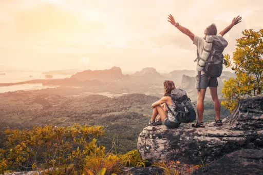Two people with backpacks sit on a rock, overlooking a scenic landscape at sunset. One person stands with arms raised.