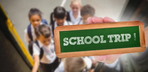 A hand holds a chalkboard sign that says SCHOOL TRIP! with children boarding a bus in the background.