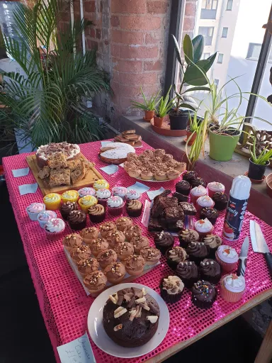 A table covered with a pink cloth displays various cakes, cupcakes, and brownies, alongside potted plants in a bright room.