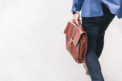 A person walking while holding a brown leather briefcase, wearing a blue blazer and dark trousers.