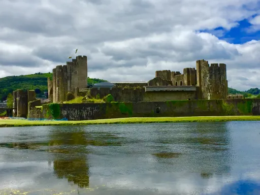 Stone castle with towers and walls reflected in a body of water under a cloudy sky.