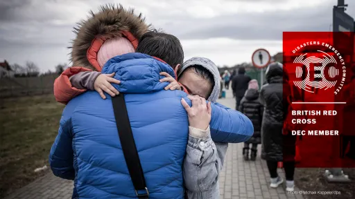 A person in a blue jacket hugs another person, while a child embraces them. People walk in the background.