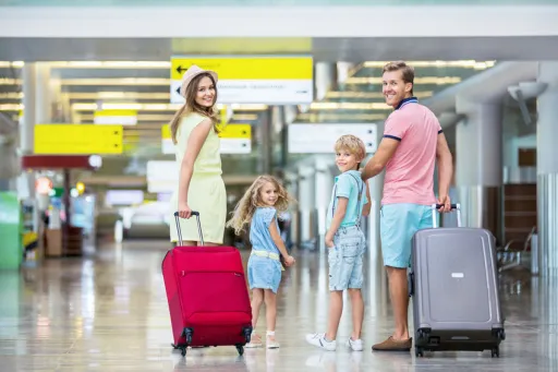 A group of four people stands in an airport terminal, smiling and holding luggage. Bright signs are visible in the backgro…