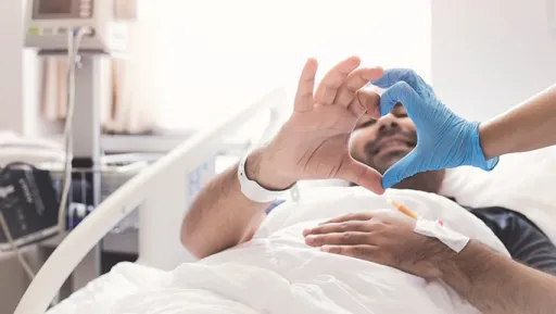 A person in a hospital bed makes a heart shape with their hands, while another hand in a blue glove reaches towards them.