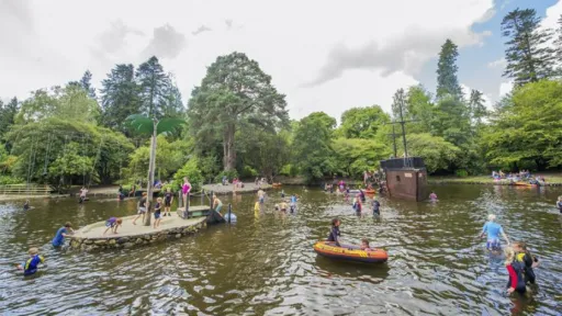 A group of people playing in a lake with a wooden pirate ship and a small island with palm trees in the background.