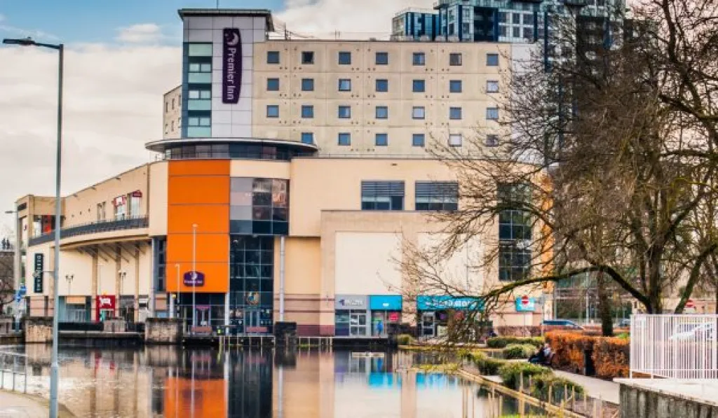 Large building with a hotel and shops reflected in a body of water, surrounded by trees.