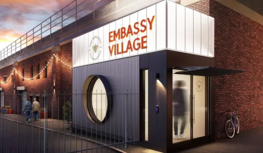 Modern building with a sign reading Embassy Village and a circular entrance, set against a brick wall.