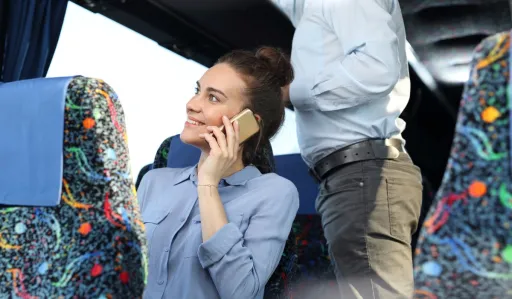 Person is sitting on a bus, smiling while talking on a mobile phone, with another person standing nearby.