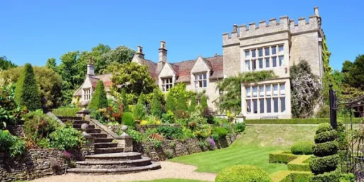 A large stone house with a garden, featuring neatly trimmed hedges, flowers, and a stone staircase leading to the entrance.