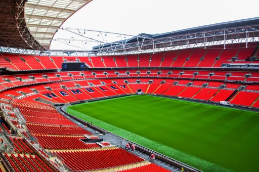 A view of Wembley Stadium, featuring red seating and a green pitch under a cloudy sky.