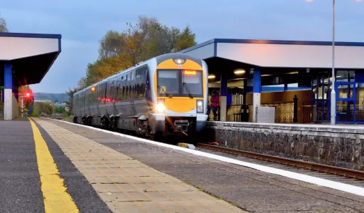 Train arrives at a platform with a station building and trees in the background under a cloudy sky.