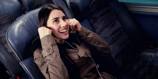 A person sitting in a bus seat, smiling and wearing headphones, with hands raised near their ears.