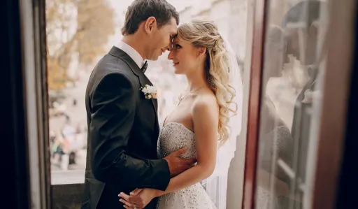 Couple stands close together, gazing into each other's eyes by a window.