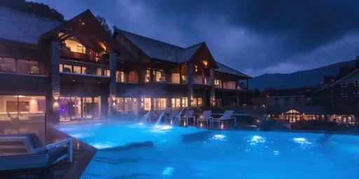 A modern building with large windows beside a blue-lit pool at night, surrounded by mist and mountains in the background.