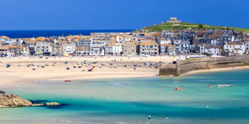 A coastal view of a beach with boats, sandy shore, and buildings along the waterfront under a clear blue sky.