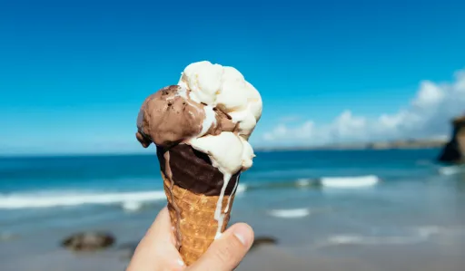 Hand holds a chocolate and vanilla ice cream cone against a beach and blue sky background.