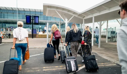 Group of people walking towards an airport terminal, pulling luggage behind them.