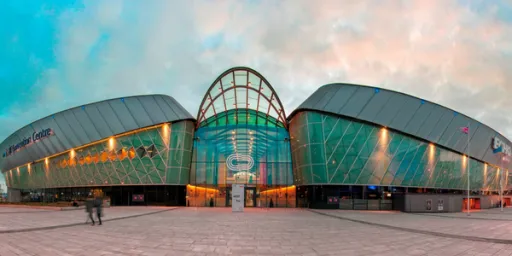 A modern building with a glass facade and a curved roof, featuring an entrance and two people walking in front.